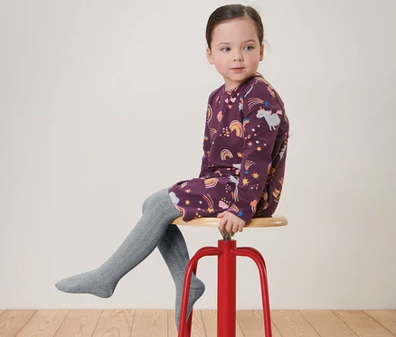 Mädchen sitzt auf einem roten Hocker und trägt ein bedrucktes Kleid und graue 2 Paar Kinder-Strumpfhosen.