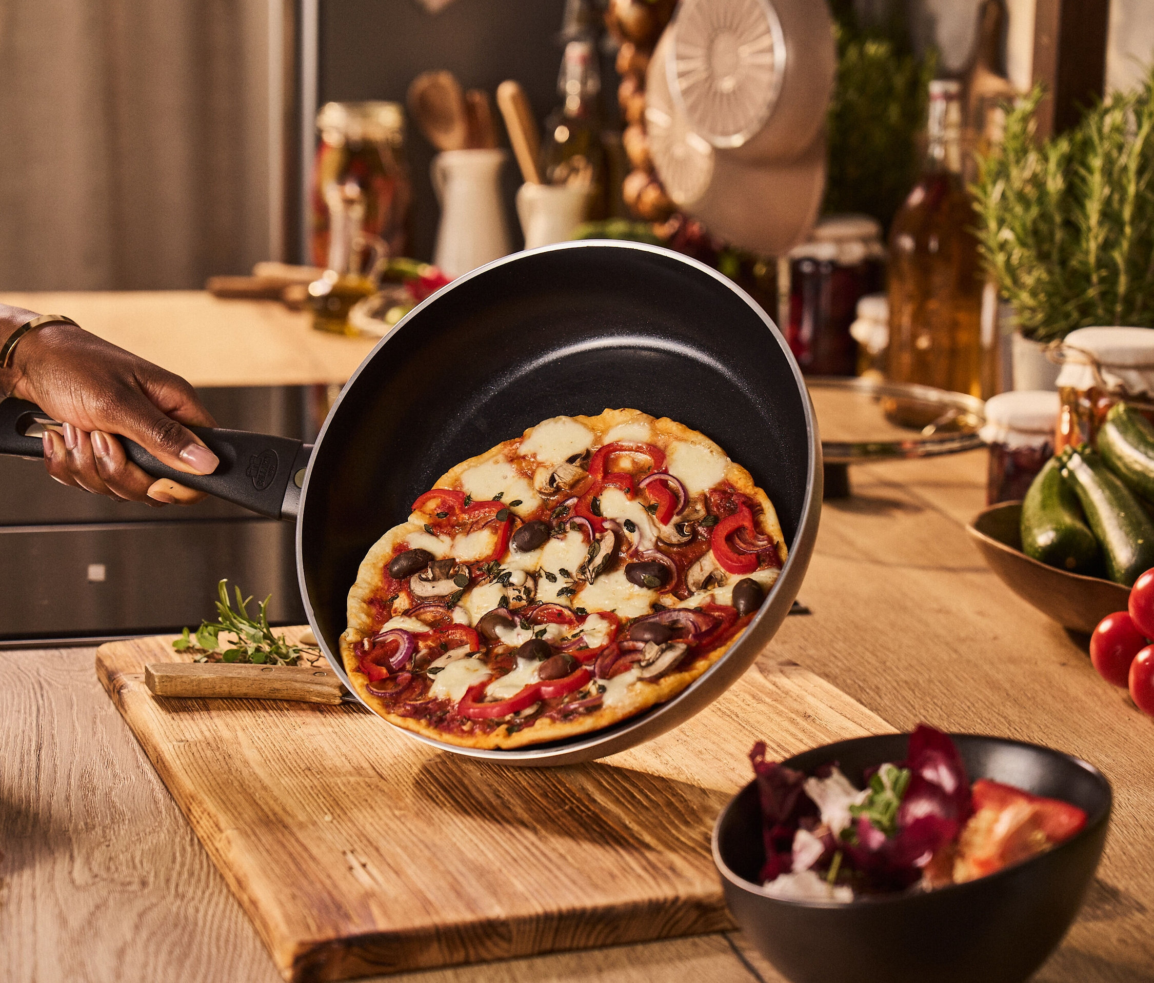 Eine Hand hält eine BALLARINI Aluminium-Bratpfanne, sand, 28 cm mit Pizza über einem Holztisch.