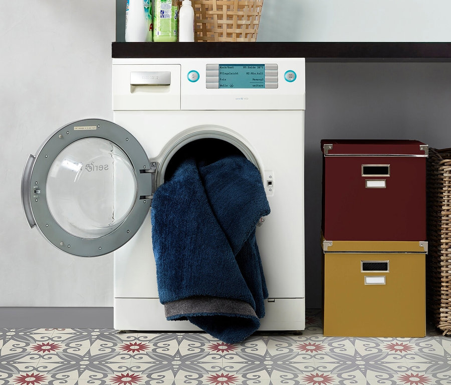 Waschmaschine mit offener Tür und blauem Handtuch im Inneren steht auf einem gemusterten Boden. Im Hintergrund sind blaue Teppiche »Mellow« zu sehen.