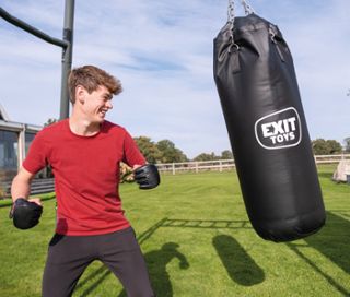 Ein junger Mann in rotem T-Shirt und schwarzen Handschuhen boxt gegen einen schwarzen EXIT Outdoor-Fitnessgerät »MB230« Boxsack auf einer grünen Wiese.