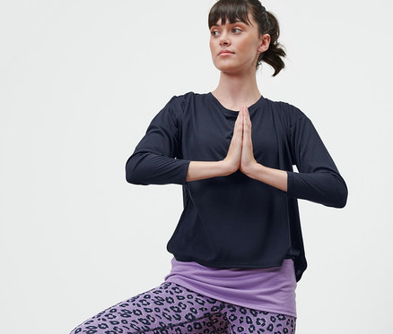 Frau im blau-lila 2-in-1-Sportshirt steht auf einem Bein in der Baum-Pose.