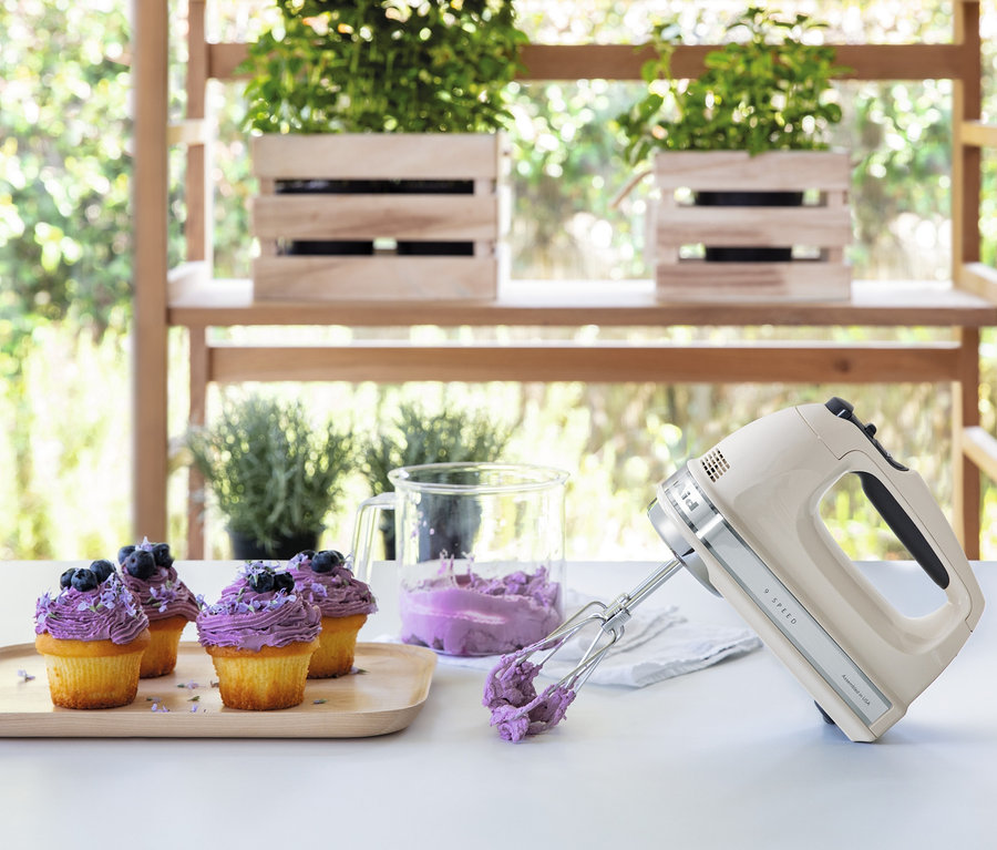 Vier Muffins mit lila Creme und Blaubeeren auf einem Holztablett neben einem KitchenAid-Handmixer »5KHM9212EAC«.