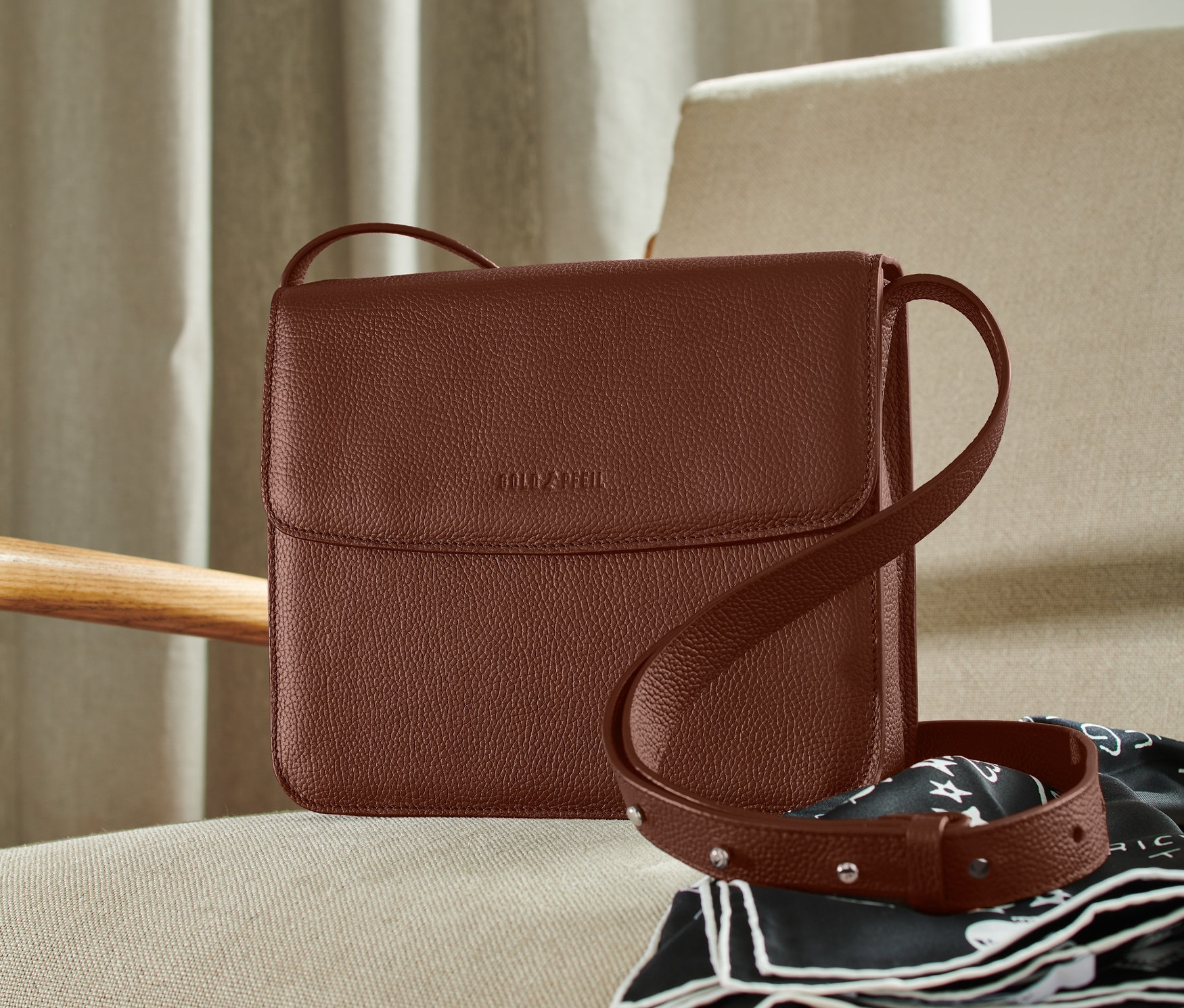 Eine braune Goldpfeil Leder-Umhängetasche liegt auf einem cremefarbenen Sessel neben einem schwarz-weißen Schal.