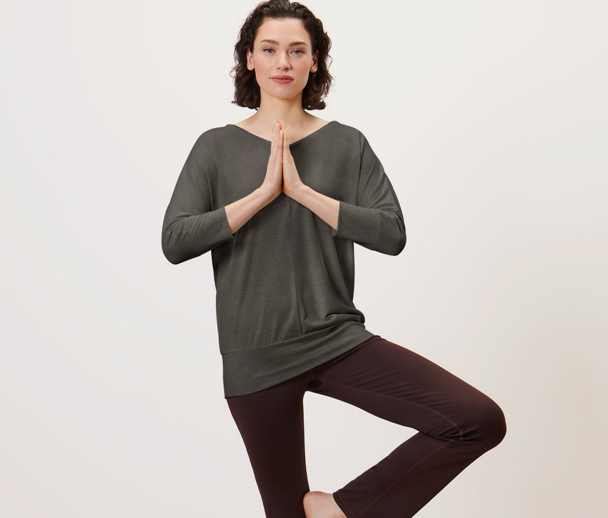 Frau im olivfarbenen Yogashirt steht auf einer Yogamatte in der Baumposition.