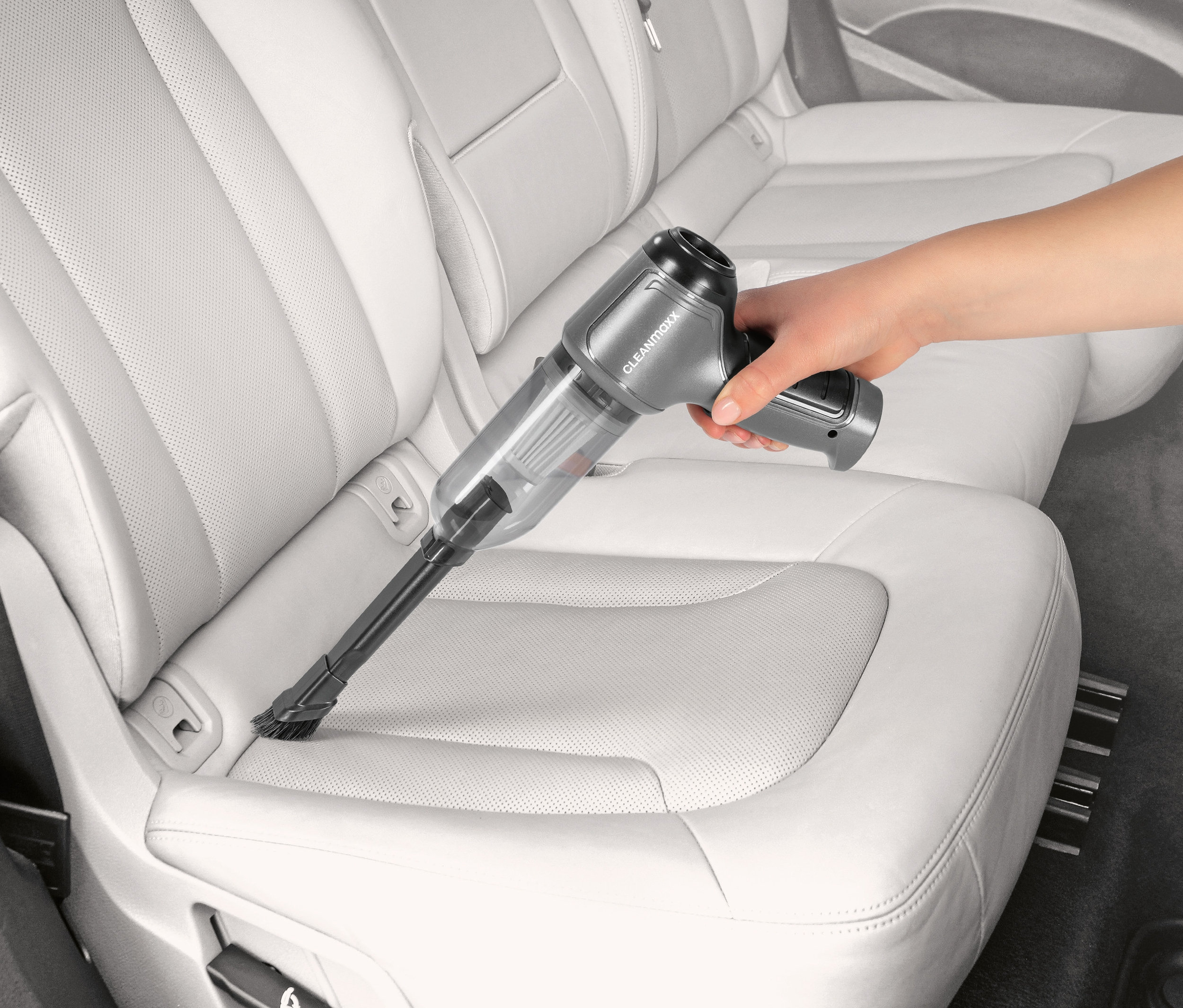 Eine Hand benutzt einen grauen Akku-Handstaubsauger, um einen weißen Ledersitz im Auto zu saugen.
