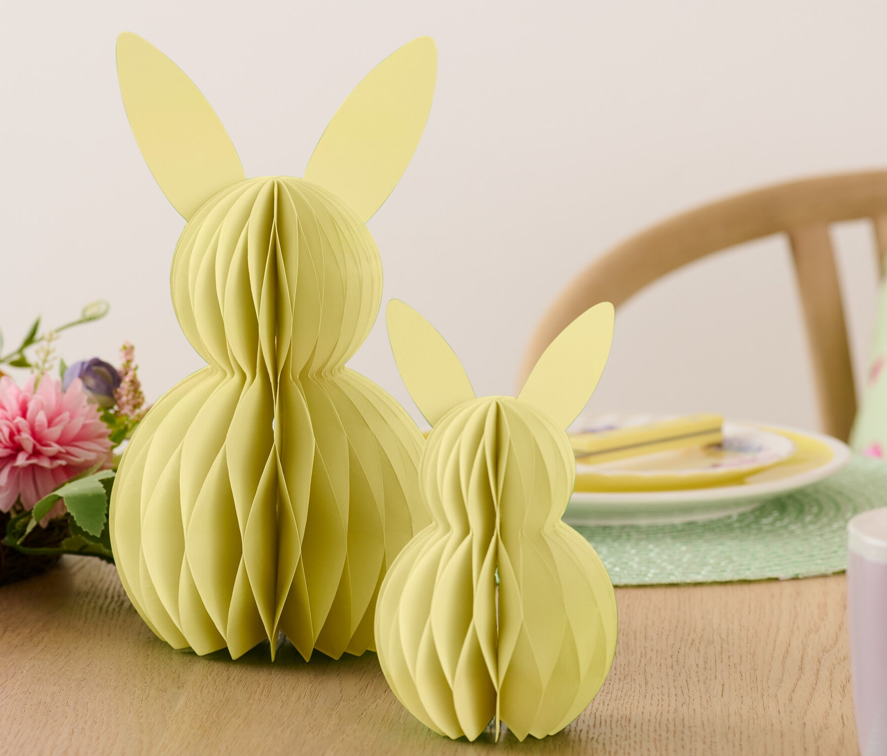 Zwei Papier-Osterhasen stehen auf einem Tisch neben Blumen und Tellern.