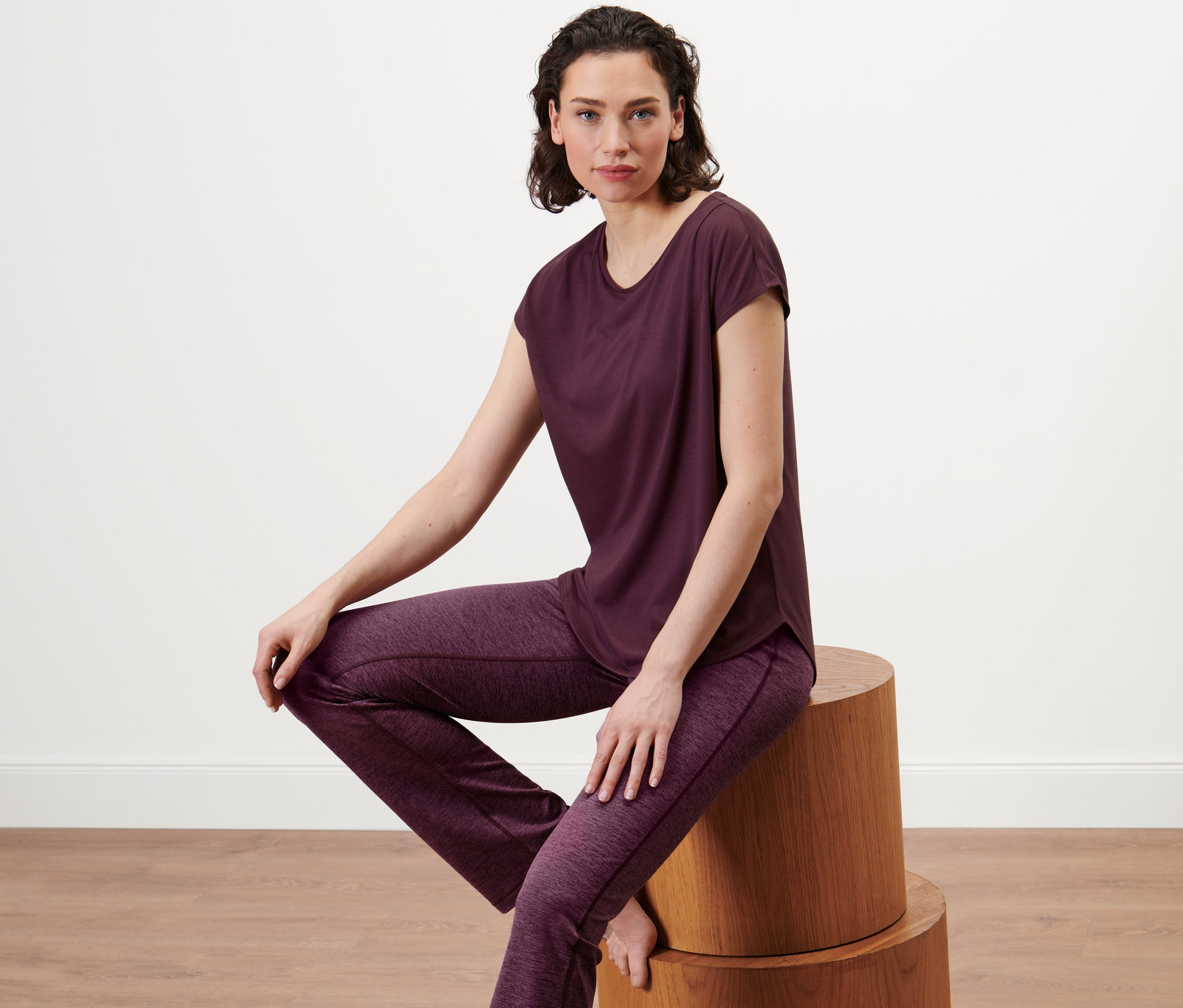 Frau sitzt auf einem Holzhocker und trägt ein Sportshirt in Pflaumenfarbe.