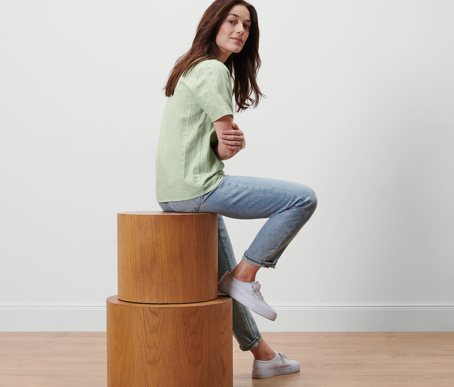 Frau sitzt auf Holzzylindern und trägt eine hellgrüne Webbluse.