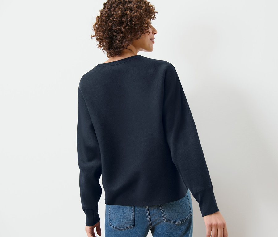 Rückansicht einer Frau mit lockigem Haar, die einen marineblauen Feinstrickpullover mit Fledermausärmeln und Jeans trägt.