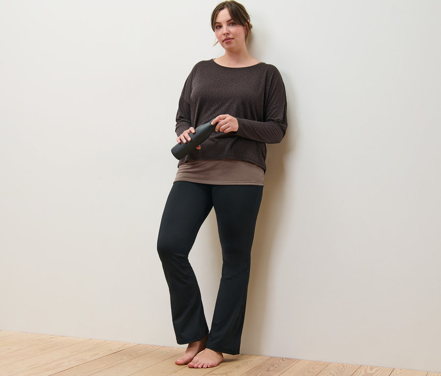 Eine Frau lehnt an einer Wand und hält ein braunes 2-in-1-Sportshirt und eine Bodyforming-Sporthose.