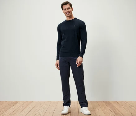 Ein Mann steht und trägt einen dunkelblauen Pullover mit Rundhalsausschnitt, dunkelblaue Hosen und weiße Turnschuhe.