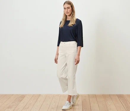 Eine Frau steht auf einem Holzboden und trägt ein marineblaues Oberteil und eine cremefarbene Flared Jeans – Fit »Cara«.