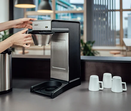 Frau setzt Filter in Profi-Filterkaffeemaschine »Bravilor-Bonamat TH« ein. Auf der Theke stehen eine Thermoskanne und drei weiße Tassen.