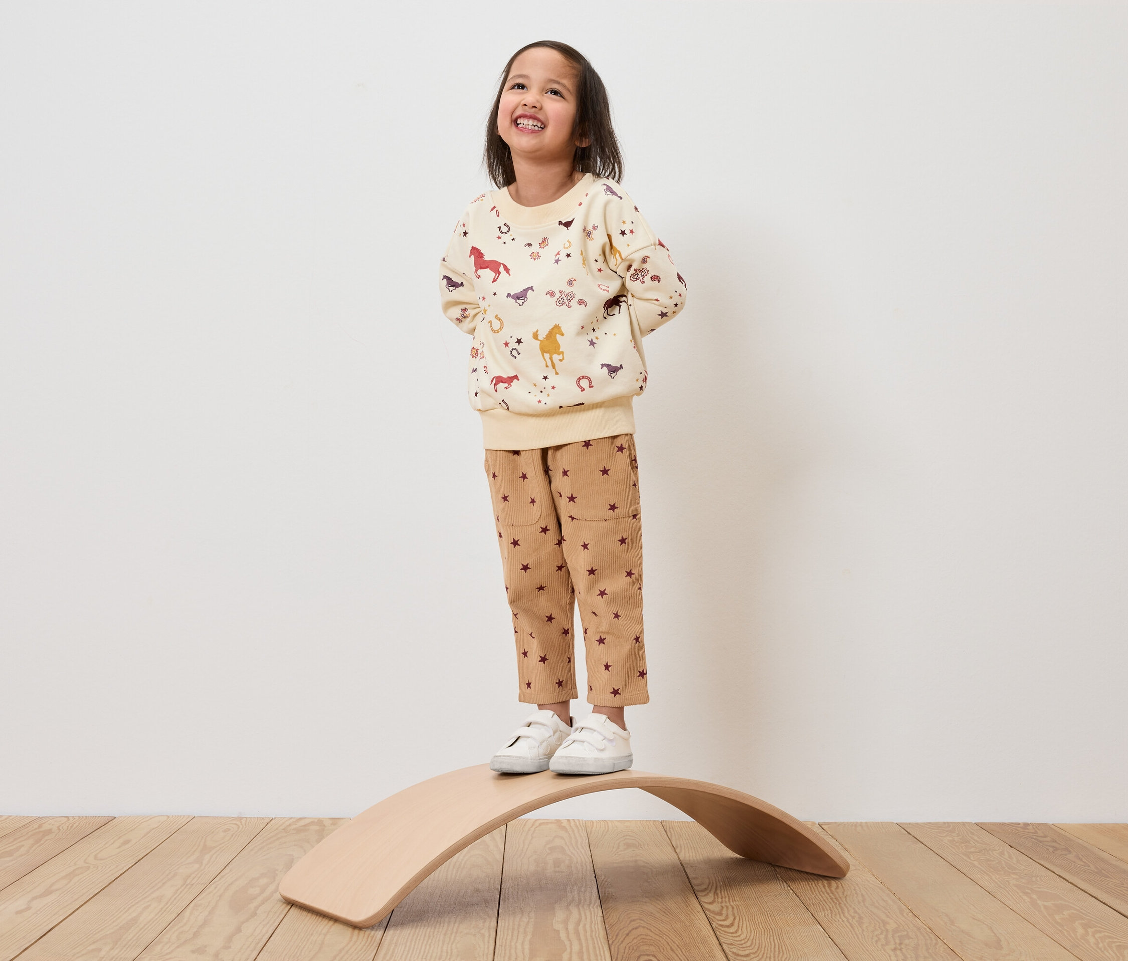Ein Mädchen steht auf einem hölzernen Balancebrett und trägt ein beiges Sweatshirt mit Pferdeaufdruck und braune Hosen mit Sternen.