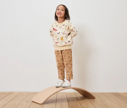 Ein Mädchen steht auf einem hölzernen Balancebrett und trägt ein beiges Sweatshirt mit Pferdeaufdruck und braune Hosen mit Sternen.