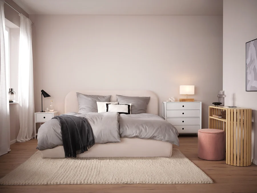 Schlafzimmer mit beigem Bett, grauer Bettwäsche und beigem Teppich. Im Hintergrund weißer Nachttisch und Kommode.