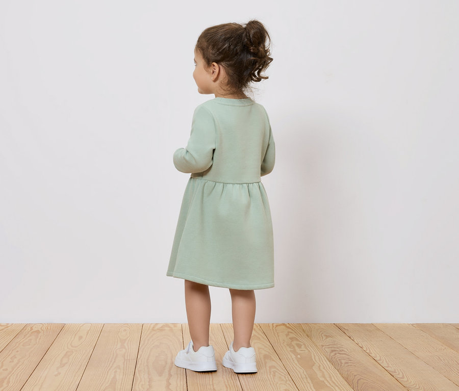 Mädchen in einem grünen Kleid und weißen Schuhen, von hinten gesehen.