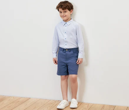 Ein Junge steht und trägt blaue Kinder-Shorts, ein Hemd und Turnschuhe.