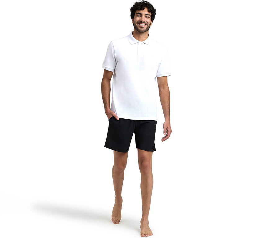 Lächelnder Mann in weißem Poloshirt und schwarzen arena Badeshorts Evo, barfuß stehend mit einer Hand in der Tasche.