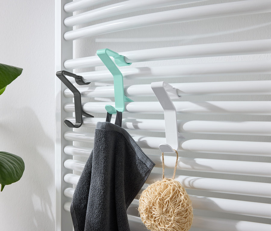 An einer weißen Heizung hängen schwarze, grüne und weiße Haken. An dem grünen Haken hängt ein graues Handtuch und an dem weißen ein Luffaschwamm.