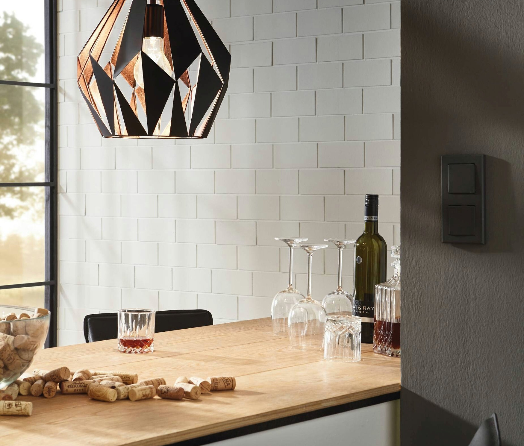 Eine schwarz-kupferfarbene, geometrische Pendelleuchte hängt über einer Bar aus Holz mit Wein, Gläsern und Korken.