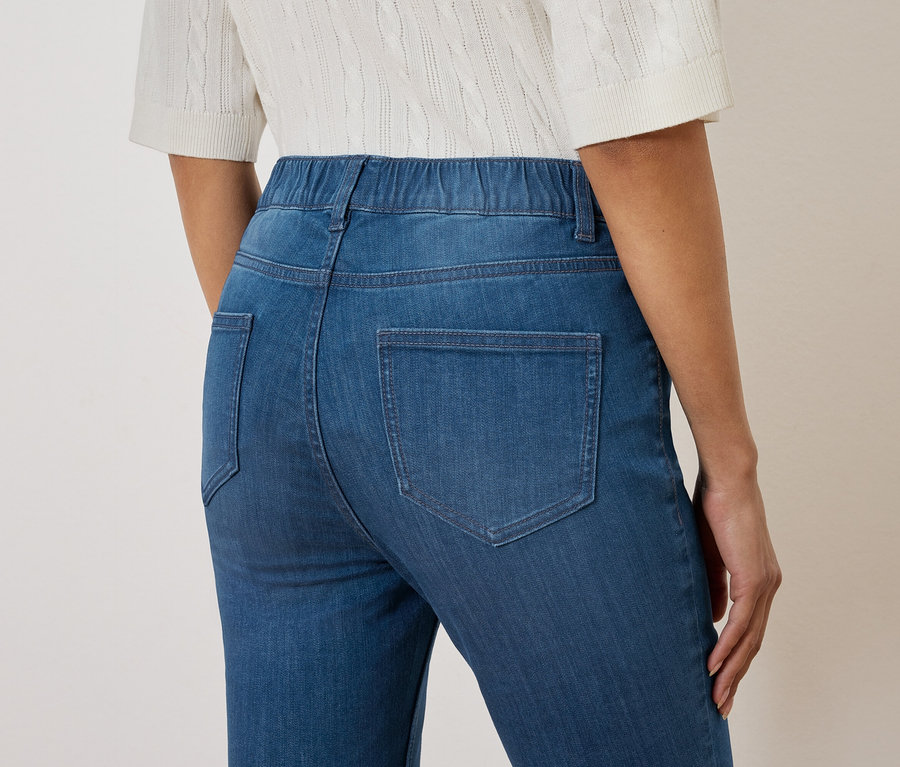 Nahaufnahme der Rückseite einer Frau, die blaue Jeans mit elastischem Bund und eine weiße Strickbluse trägt.