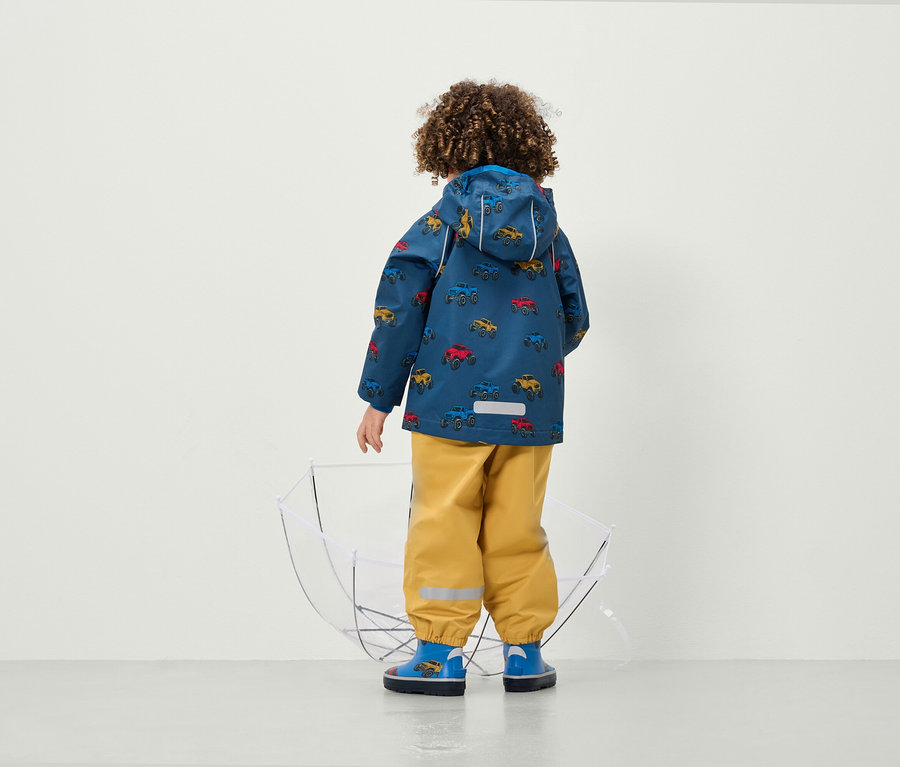 Junge mit dem Rücken zur Kamera trägt eine Jacke mit Autoaufdruck und gelbe Hosen neben einem durchsichtigen Regenschirm.