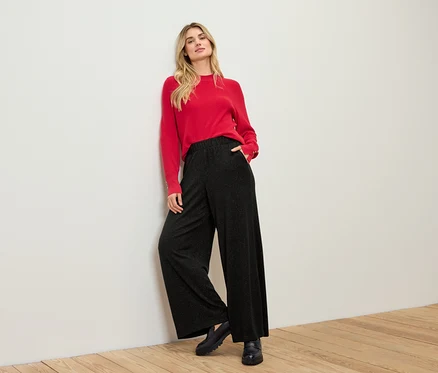 Blonde Frau in rotem Pullover und schwarzer Hose lehnt an einer weißen Wand.