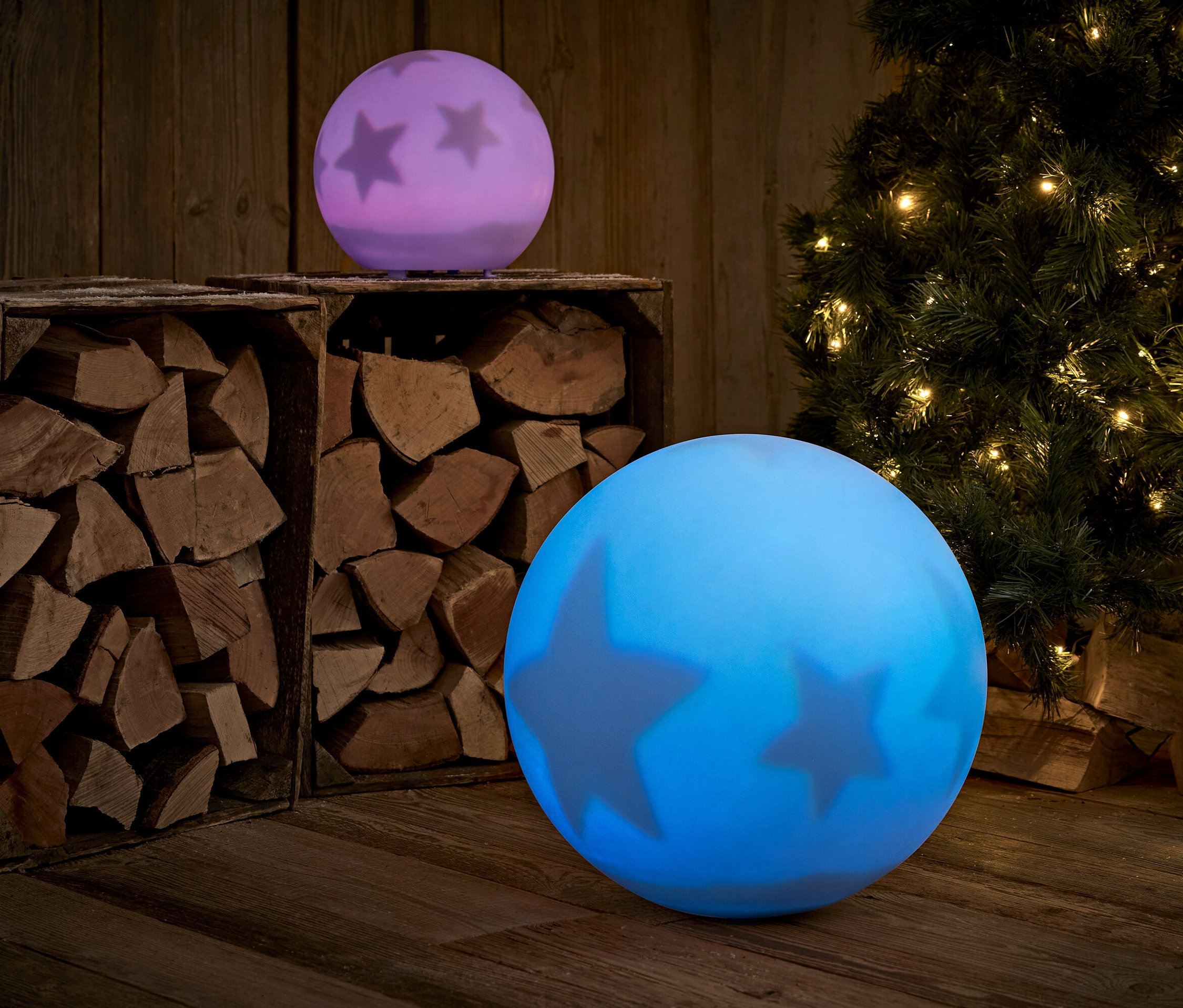 Zwei leuchtende Kugeln mit Sternen stehen auf einem Holzboden, im Hintergrund Holz und ein Weihnachtsbaum.