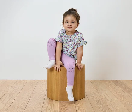Mädchen sitzt auf einem Holzhocker. Sie trägt ein geblümtes T-Shirt und lila Leggings mit Katzenmotiv.