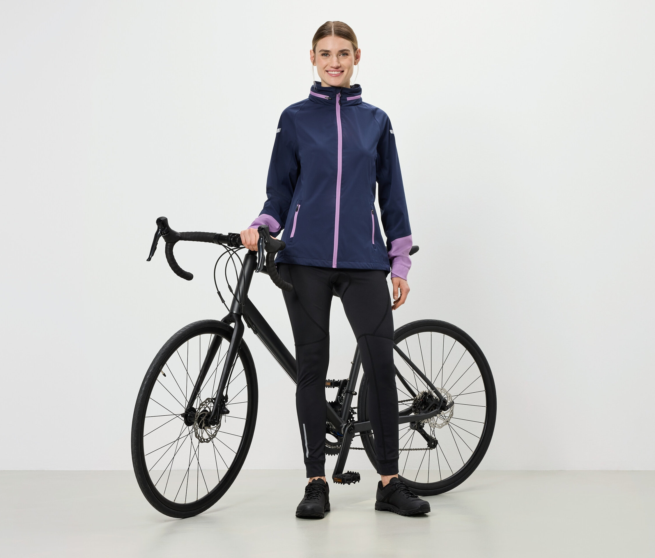 Eine Frau steht neben einem Fahrrad, sie trägt eine Jacke mit violetten Akzenten, schwarze Sportleggings und Schuhe.