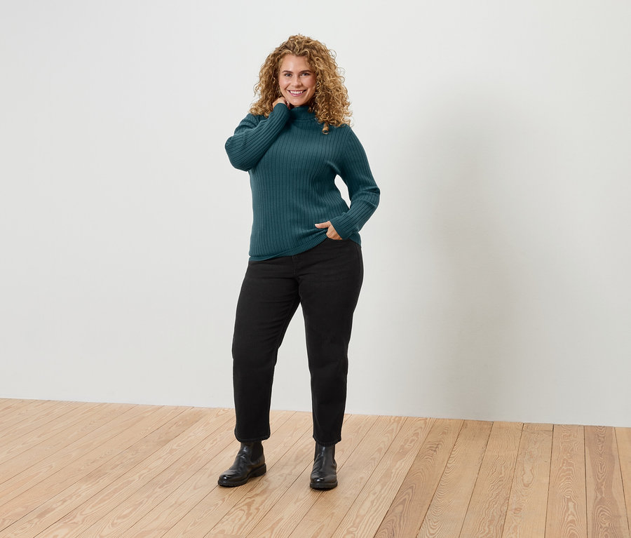 Frau mit lockigem Haar posiert in einem grünen Rollkragenpullover, schwarzen Hosen und Stiefeln.