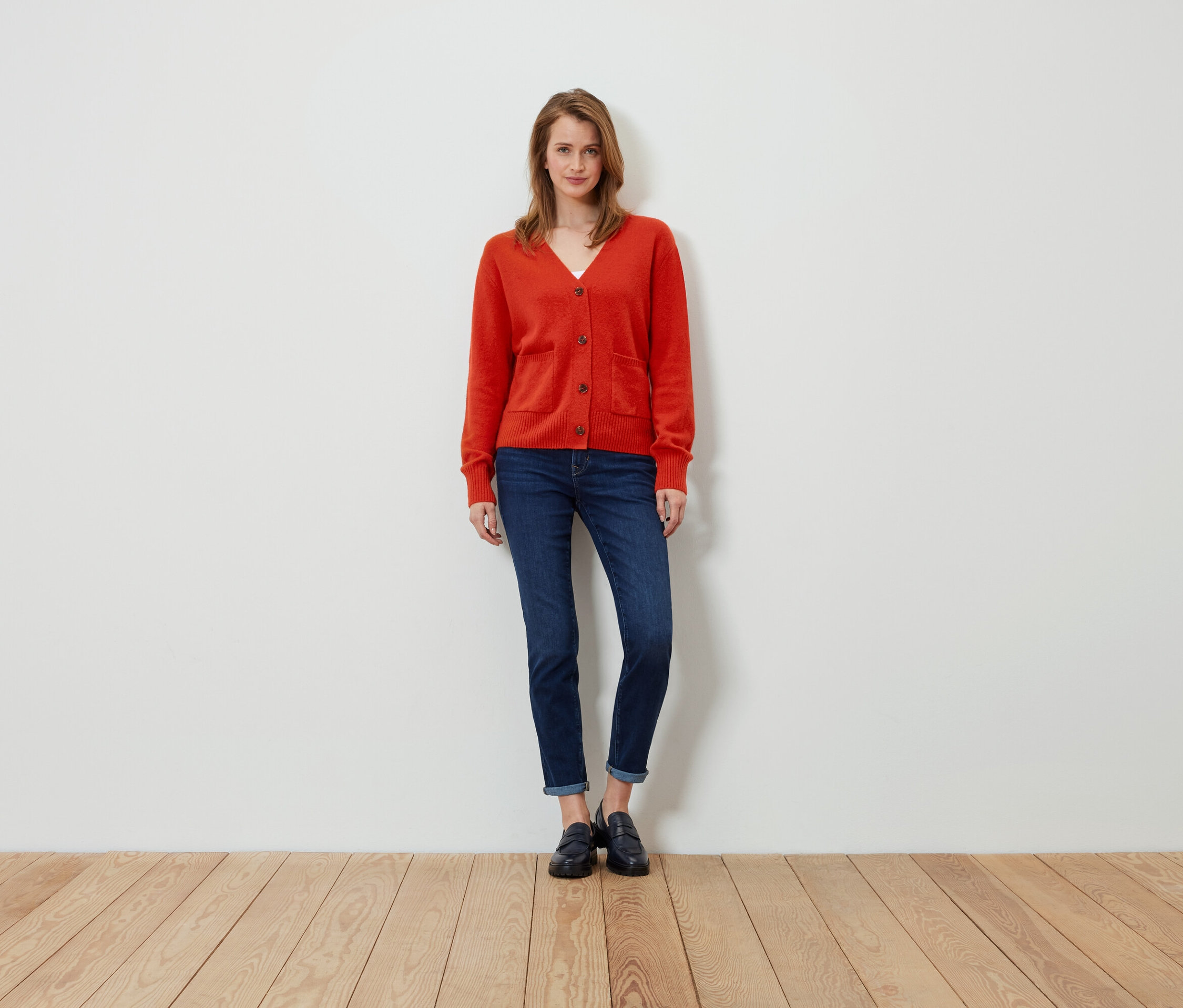 Eine Frau steht und trägt eine orangefarbene Strickjacke, Jeans und schwarze Slipper.