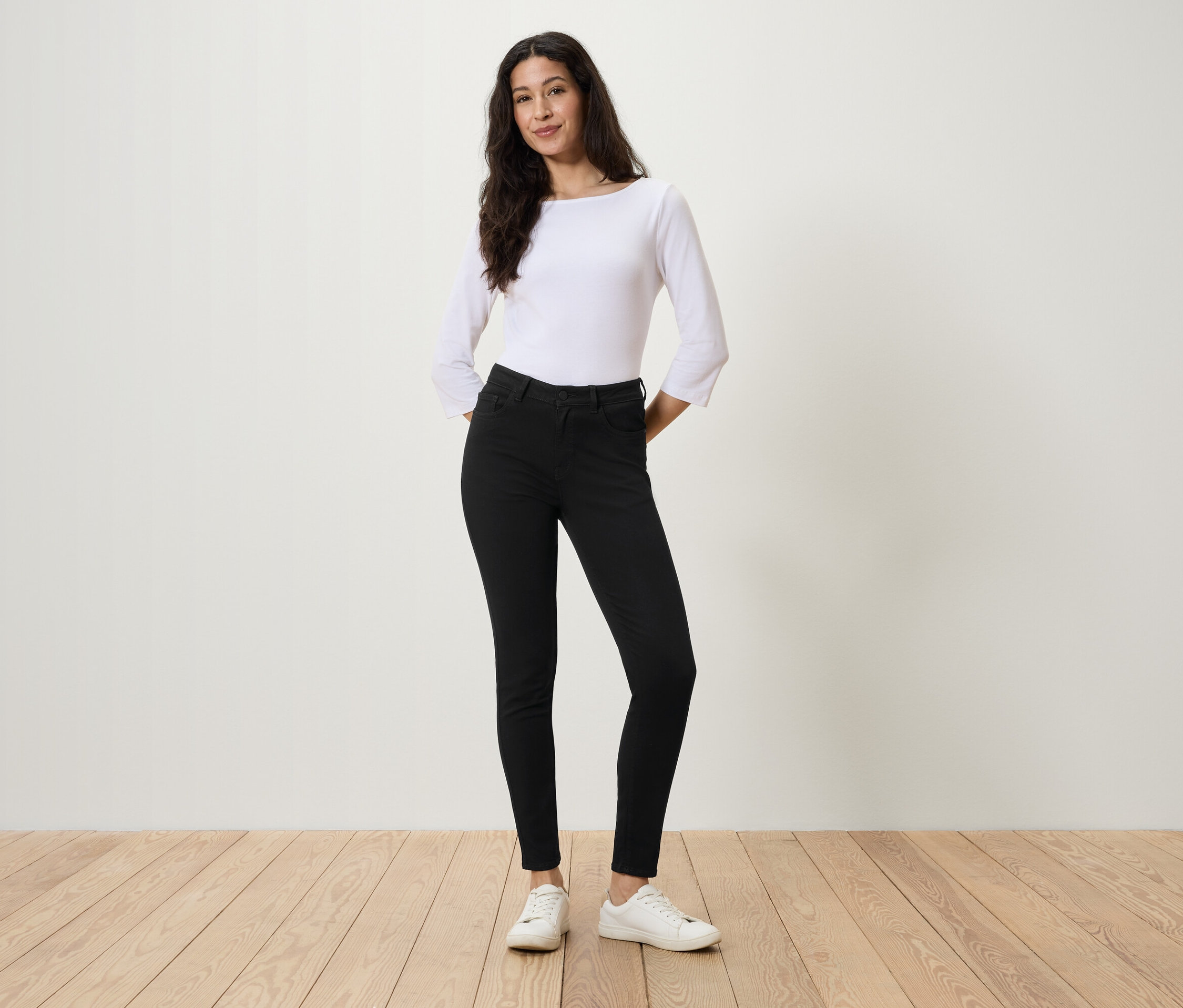 Frau steht mit Händen in den Taschen schwarzer Jeans, weißer Bluse und weißen Turnschuhen.