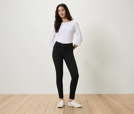 Frau steht mit Händen in den Taschen schwarzer Jeans, weißer Bluse und weißen Turnschuhen.