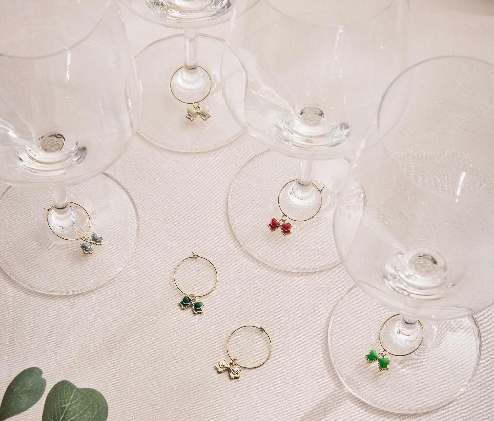 Sechs leere Weingläser mit farbigen Schleifen an den Stielen auf einer hellen Oberfläche mit einem Pflanzenblatt.