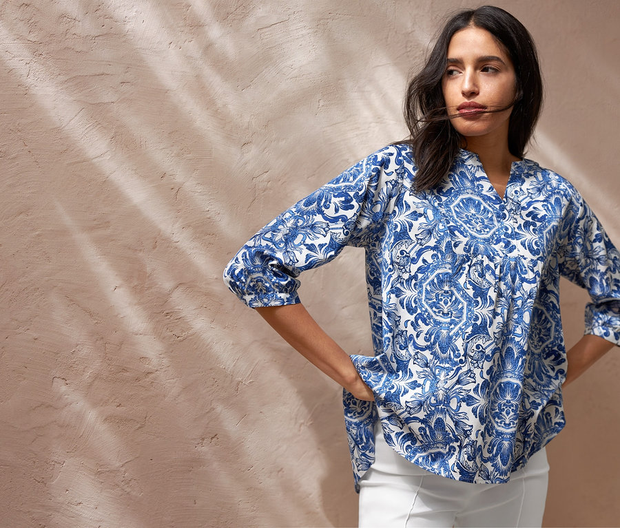 Eine Frau posiert in einer bedruckten Tunika-Bluse, einer weißen 7/8-Stretchhose und Canvas-Sneakern. Sie hat eine Umhängetasche.