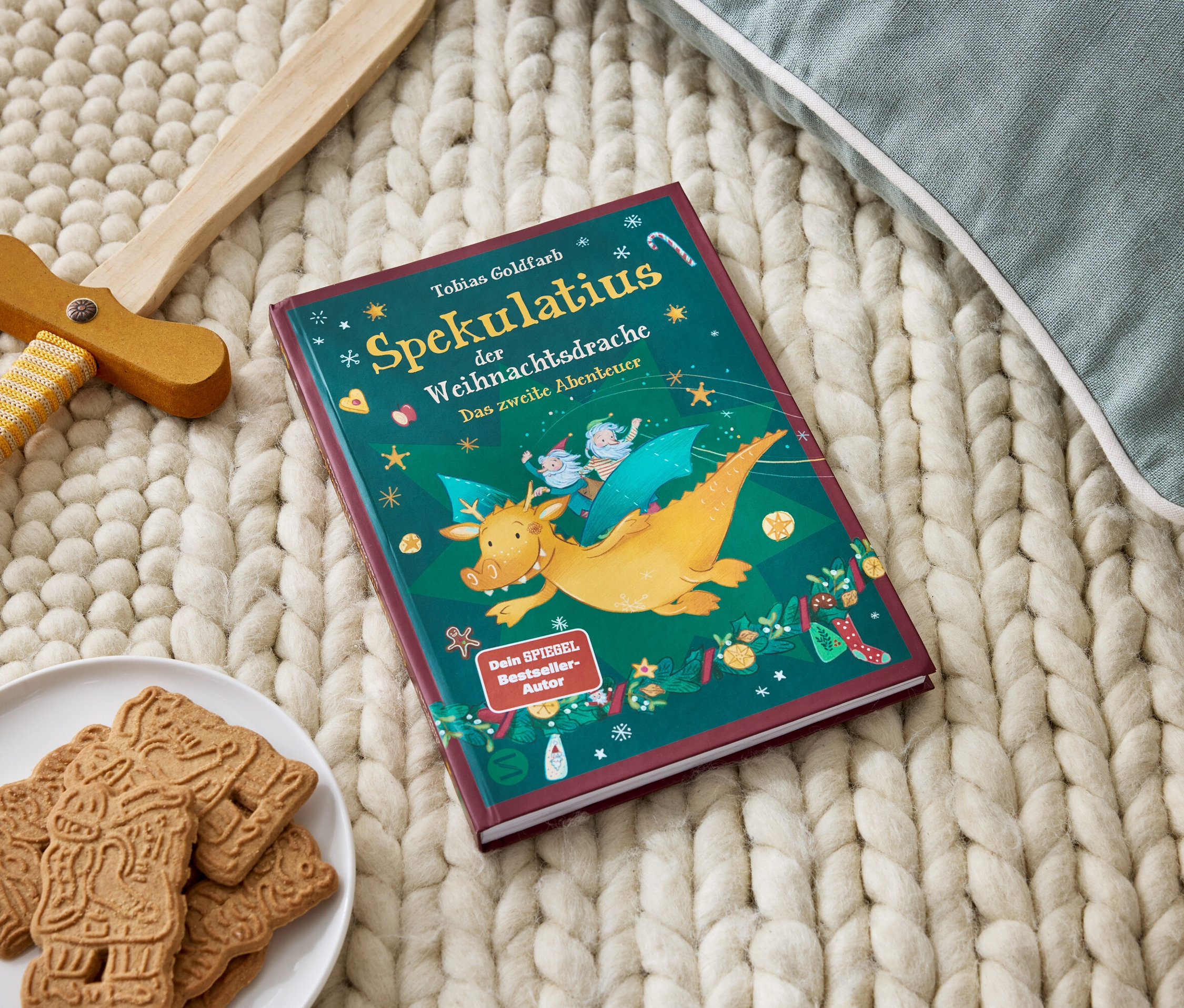 Buch Spekulatius der Weihnachtdsrache liegt auf einer Strickdecke neben einem Teller mit Lebkuchen und einem Holzschwert.
