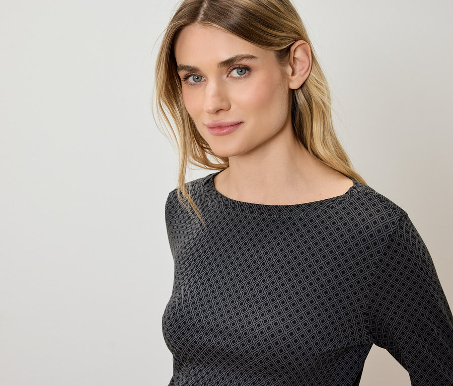Porträt einer Frau mit blonden Haaren und blauen Augen in einem grau-schwarzen Oberteil mit geometrischem Muster.