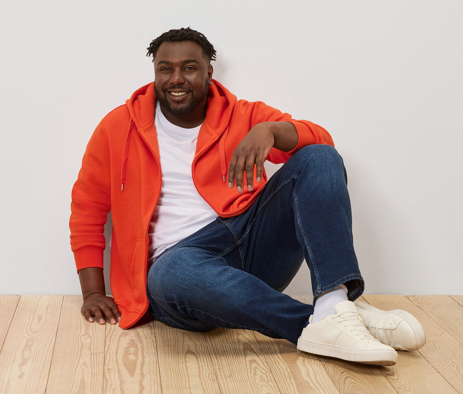 Mann sitzt auf Holzboden und trägt eine orange Kapuzenjacke.