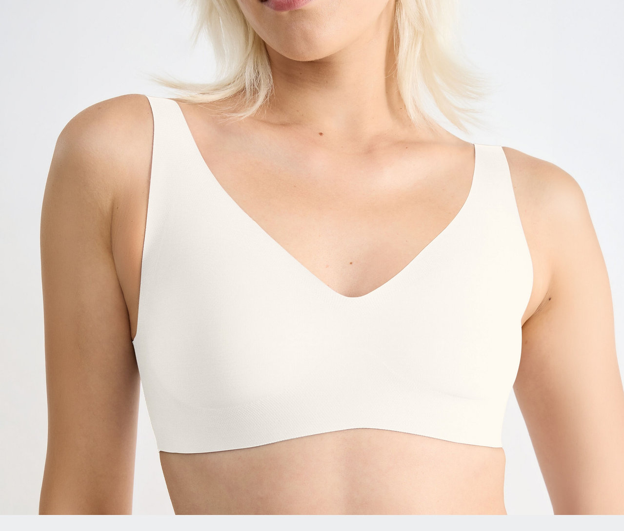 Blonde Frau trägt einen seidenweißen sloggi ZERO Feel 2.0 Soft Bra mit V-Ausschnitt, Brustbereich sichtbar.