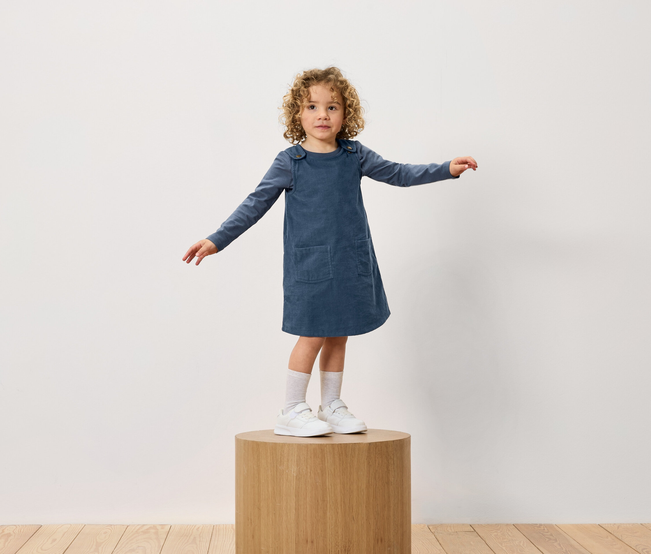 Ein Mädchen mit lockigem Haar steht auf einem Holzsockel. Sie trägt ein blaues Kleid und weiße Turnschuhe.