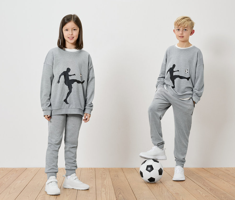 Zwei Kinder tragen graue Trainingsanzüge mit Fußballmotiven. Ein Junge steht mit einem Fuß auf einem Fußball.
