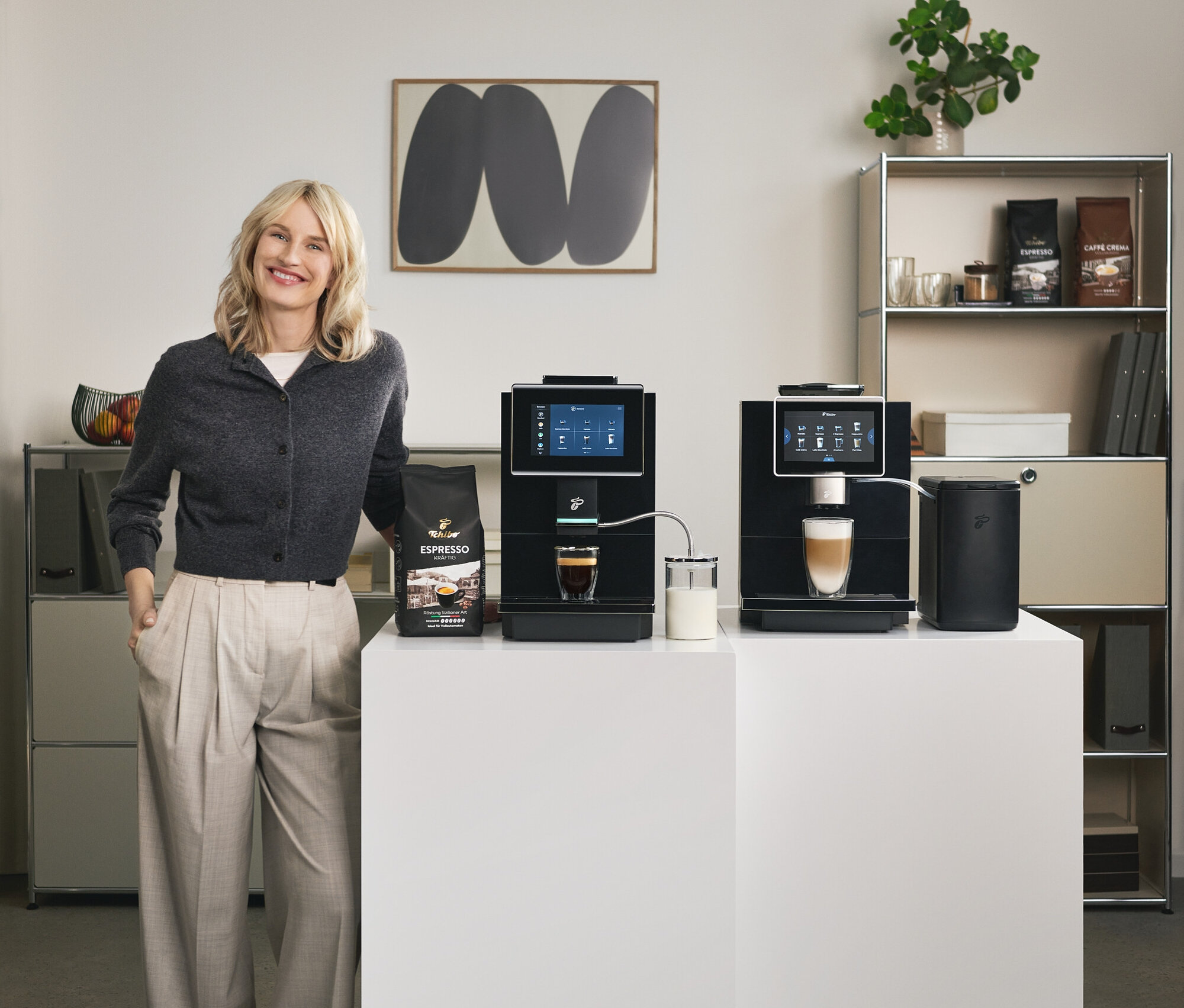 Eine lächelnde Frau steht neben zwei Tchibo Kaffeemaschinen auf weißen Podesten. Auf dem linken Podest steht eine Packung Tchibo Espresso. Eine Tasse Espresso steht unter der linken Maschine.