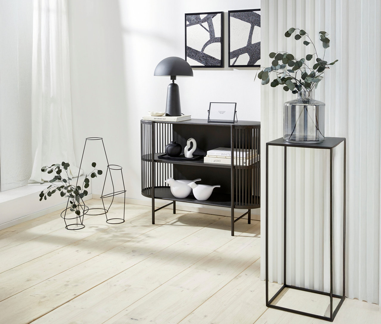 Dekorativer Raum mit einem schwarzen Regal, einer Lampe, Kunstwerken und einer Blumenvase auf einem Sockel.