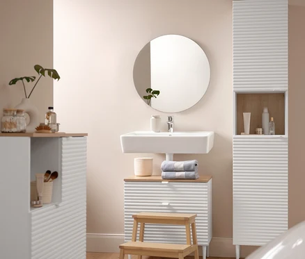 Ein Badezimmer zeigt einen Badhochschrank »SONARI« mit Türen, einen Waschbeckenunterschrank »SONARI« mit Schubladen und eine Badkommode »SONARI« mit Türen. Eine Komfort-Badematte (60 x 100 cm) liegt vor einem Tritthocker aus Massivholz. Auf dem Waschbeckenunterschrank liegen zwei blaue Premium-Handtücher. Ein runder Spiegel »Ameca« hängt über dem Waschbecken.