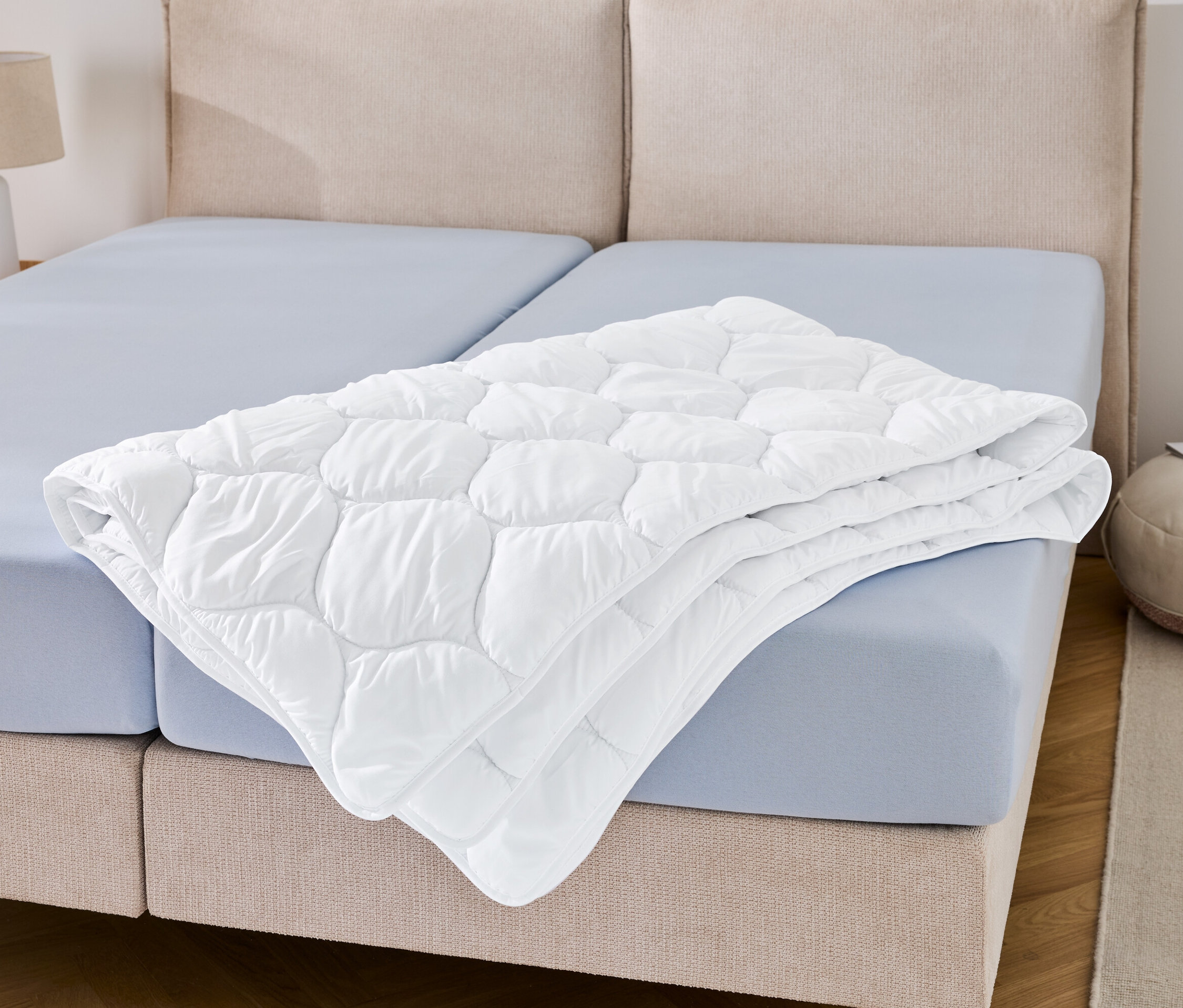 Eine kochfeste Steppdecke in Übergröße liegt gefaltet auf einem blauen Bettlaken.