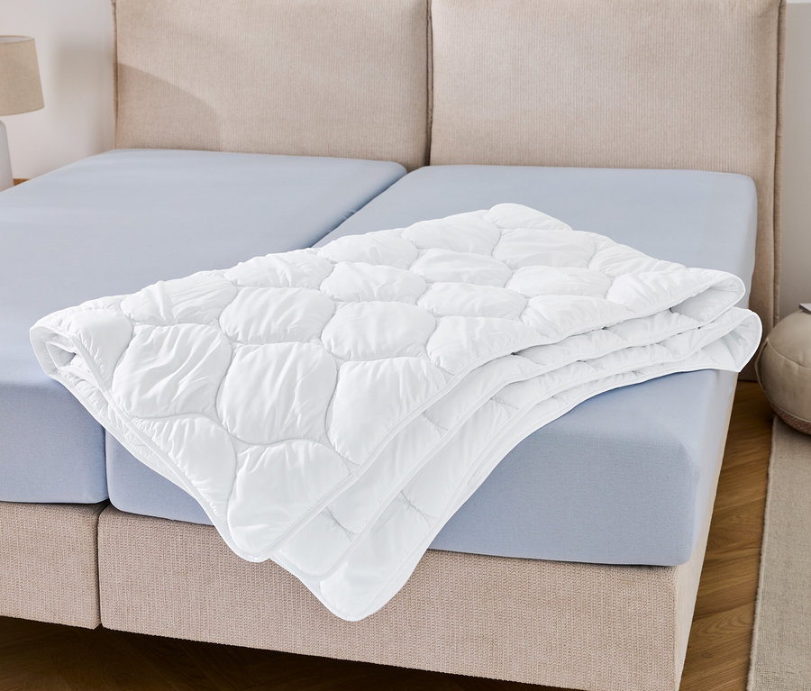 Eine kochfeste Steppdecke in Übergröße liegt gefaltet auf einem blauen Bettlaken.