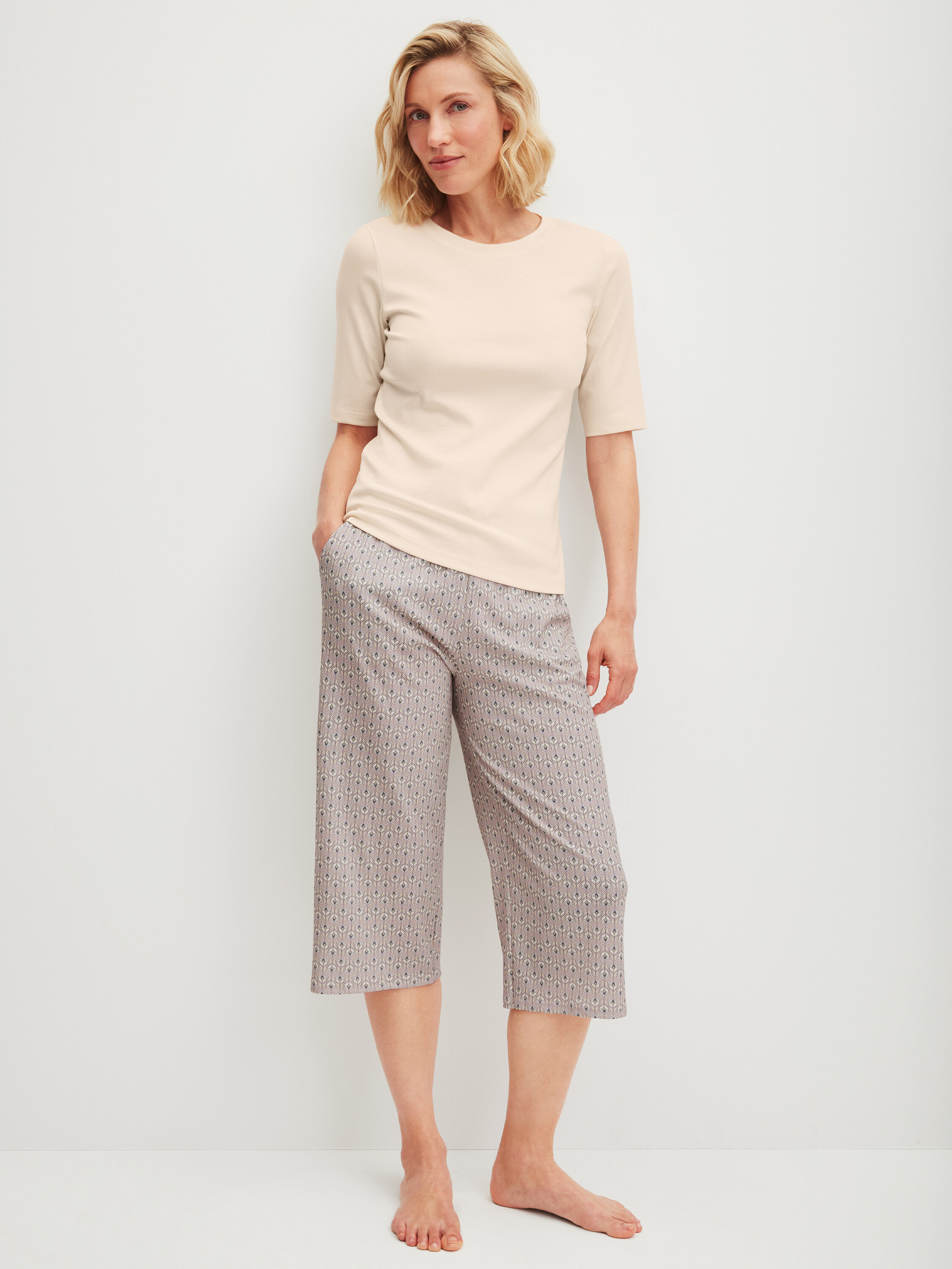 Frau steht barfuß, trägt ein helles T-Shirt und gemusterte Dreiviertelhosen.