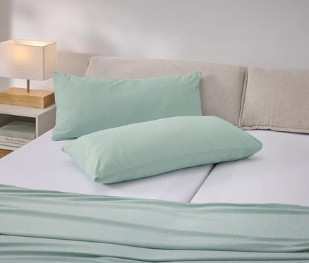 Bett mit hellgrünen Kissen und Tagesdecke auf weißer Bettwäsche. Nachttisch mit Lampe links.